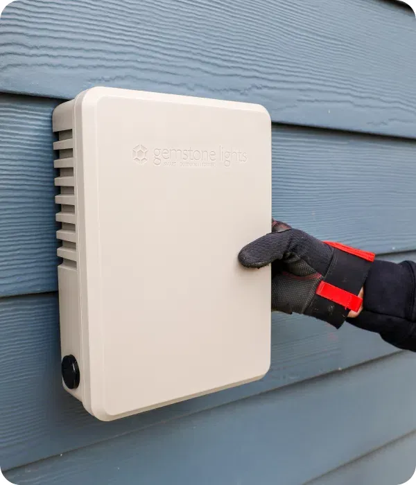 Gloved hand next to a white Gemstone Lights control box mounted on gray siding