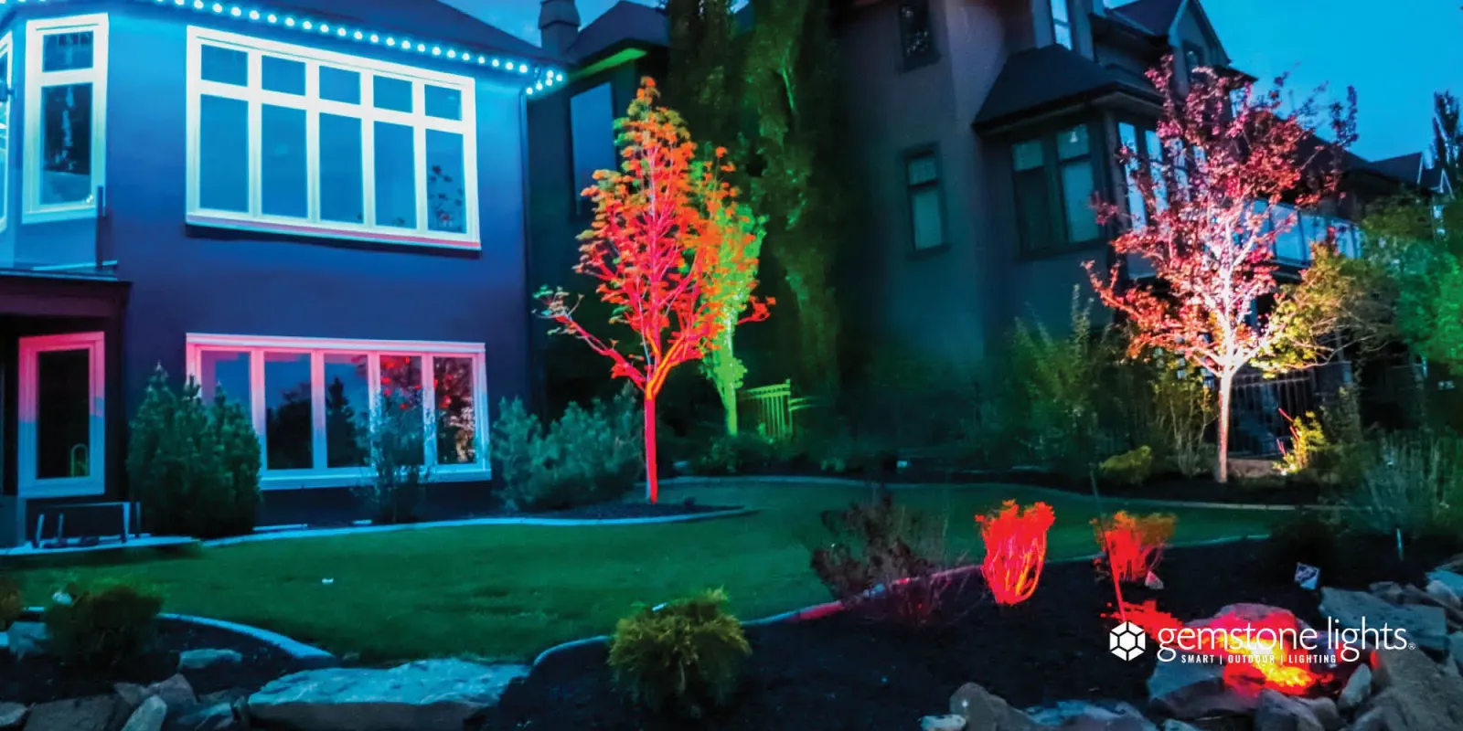 Two front-yard trees lit red and pink by smart flood lights beside a lit two-story house