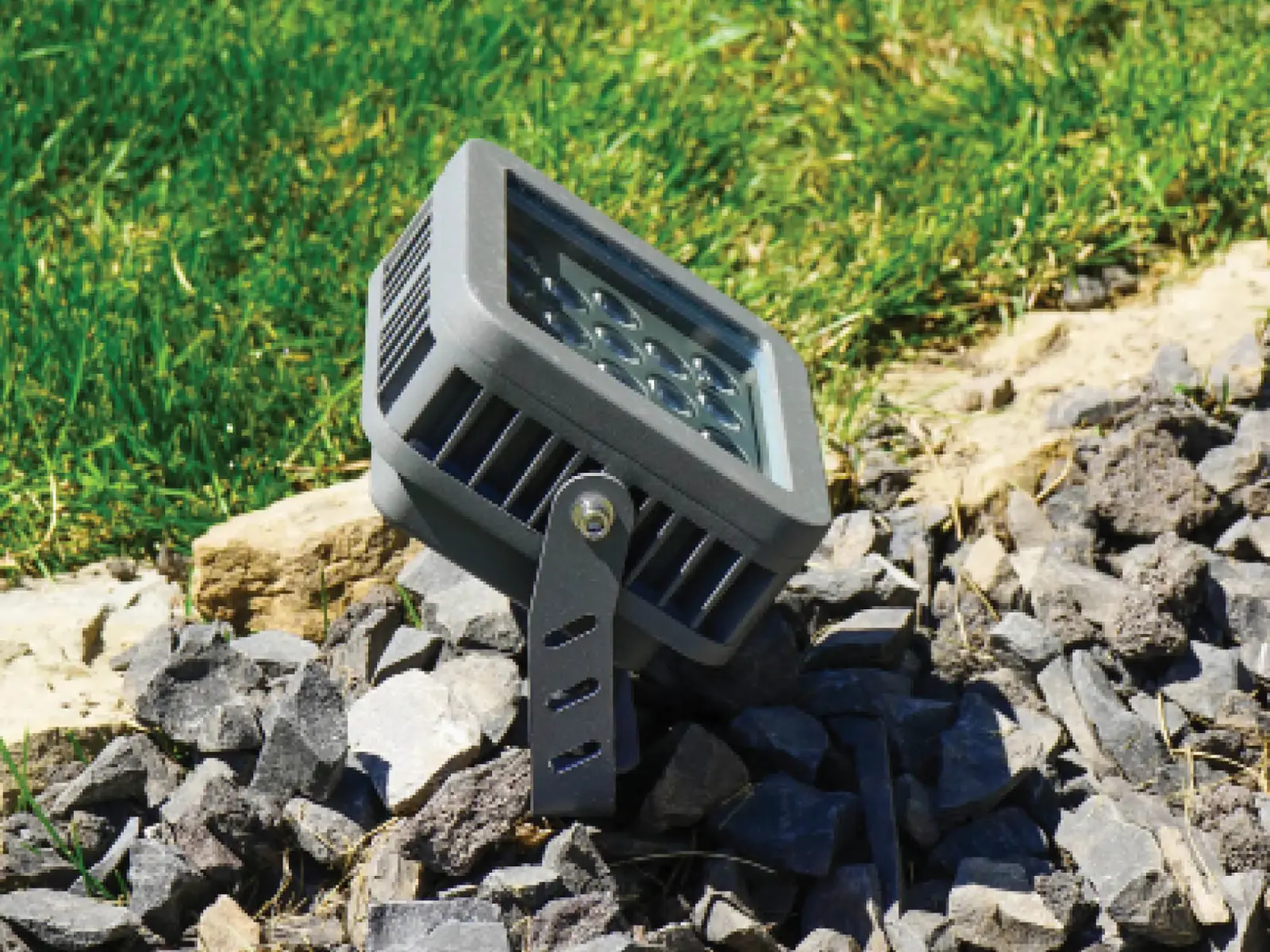 Gemstone flood light fixture angled up from a bed of landscape rock beside a lawn