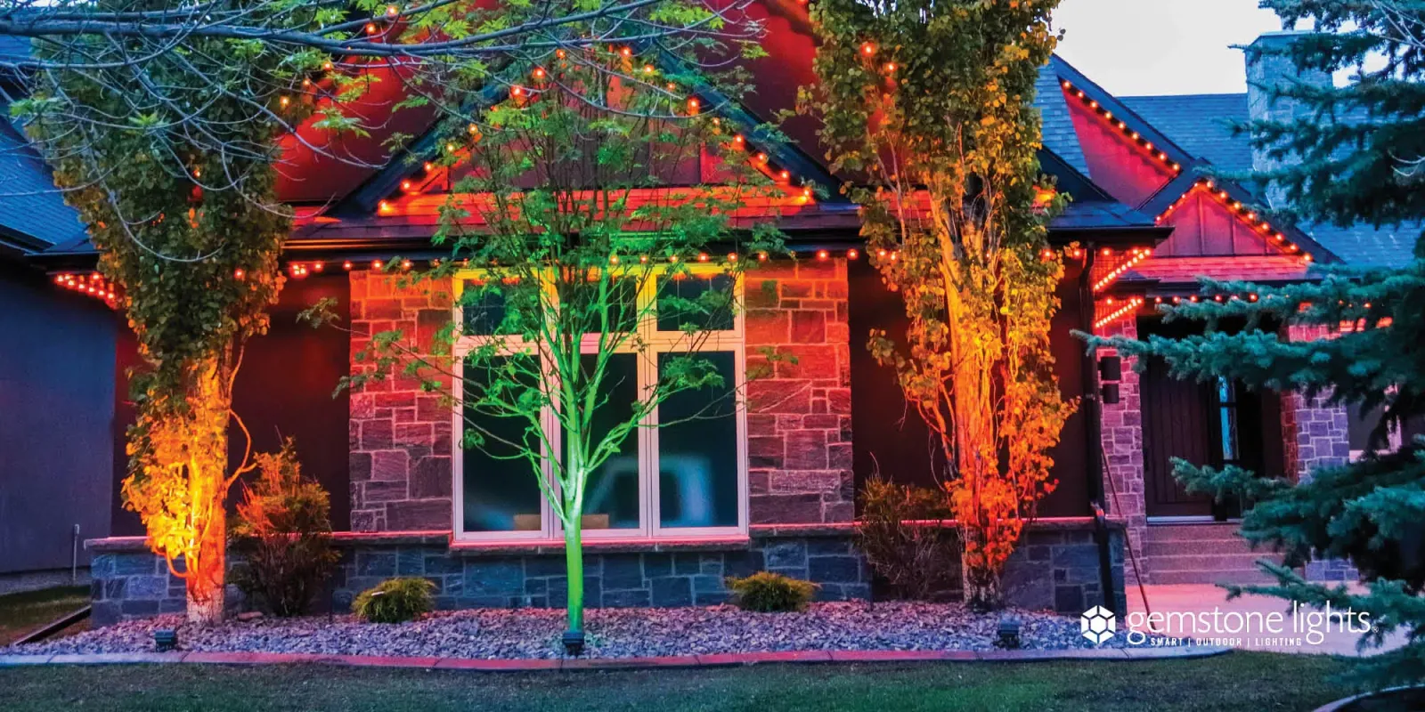 Brick home with front-yard trees lit by colored flood lights