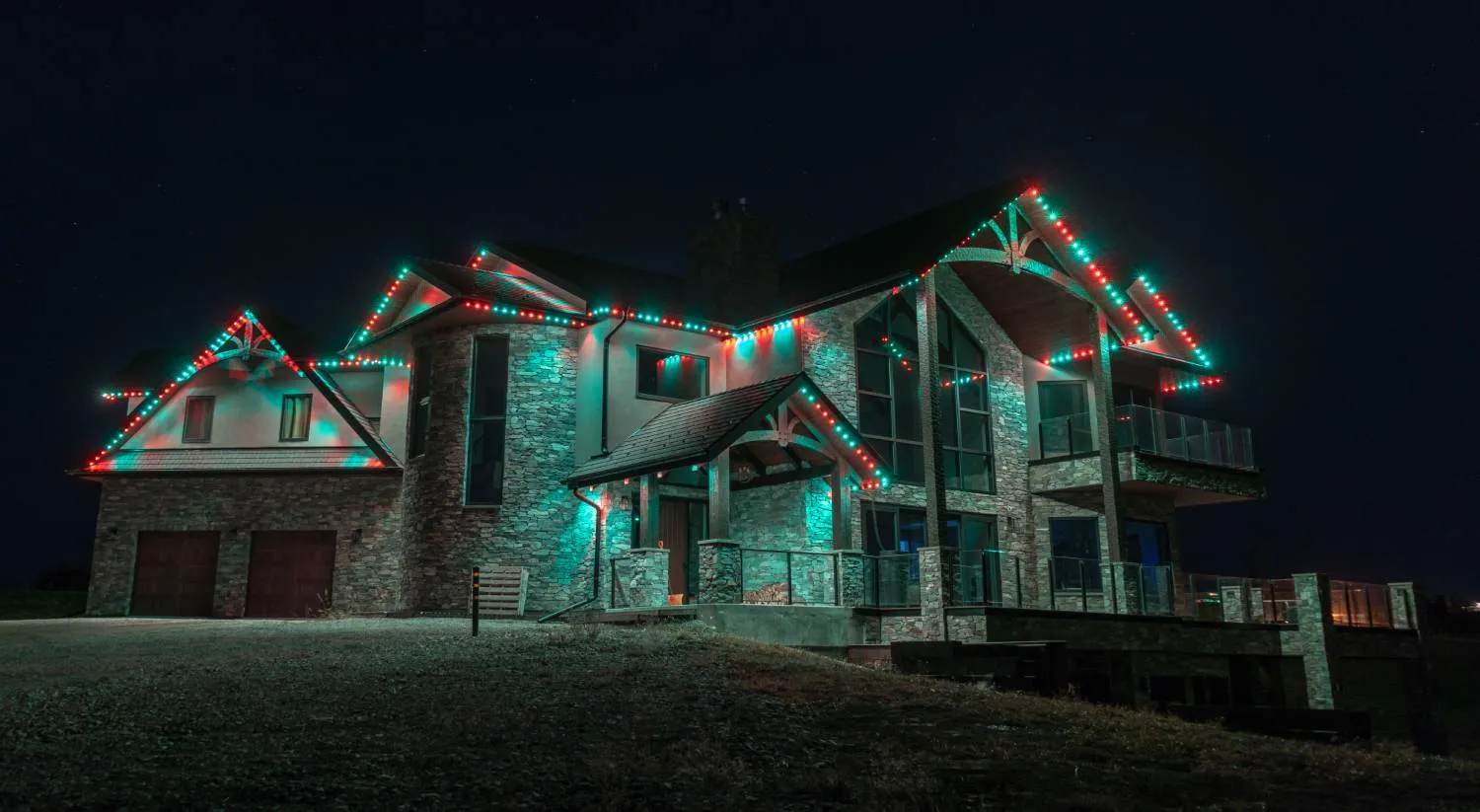 Large stone and timber home at night with red and teal holiday colors along its gables