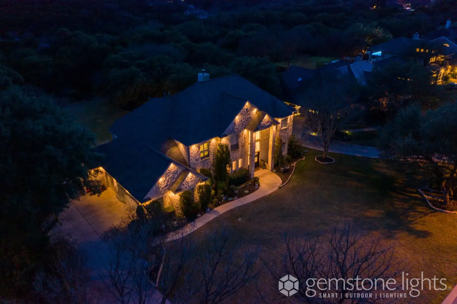 Aerial view of a home on a wooded hillside at night, its rooflines outlined in warm white lights