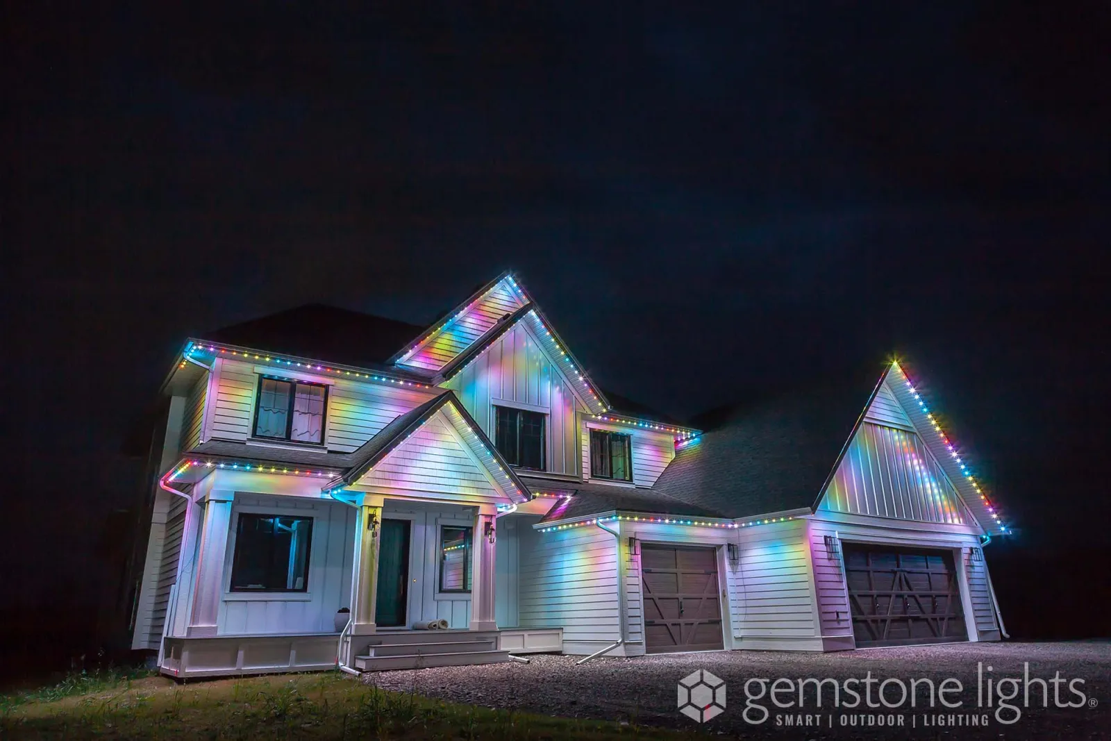 White farmhouse-style home with board-and-batten gables lit in alternating multicolor lights under a dusky sky