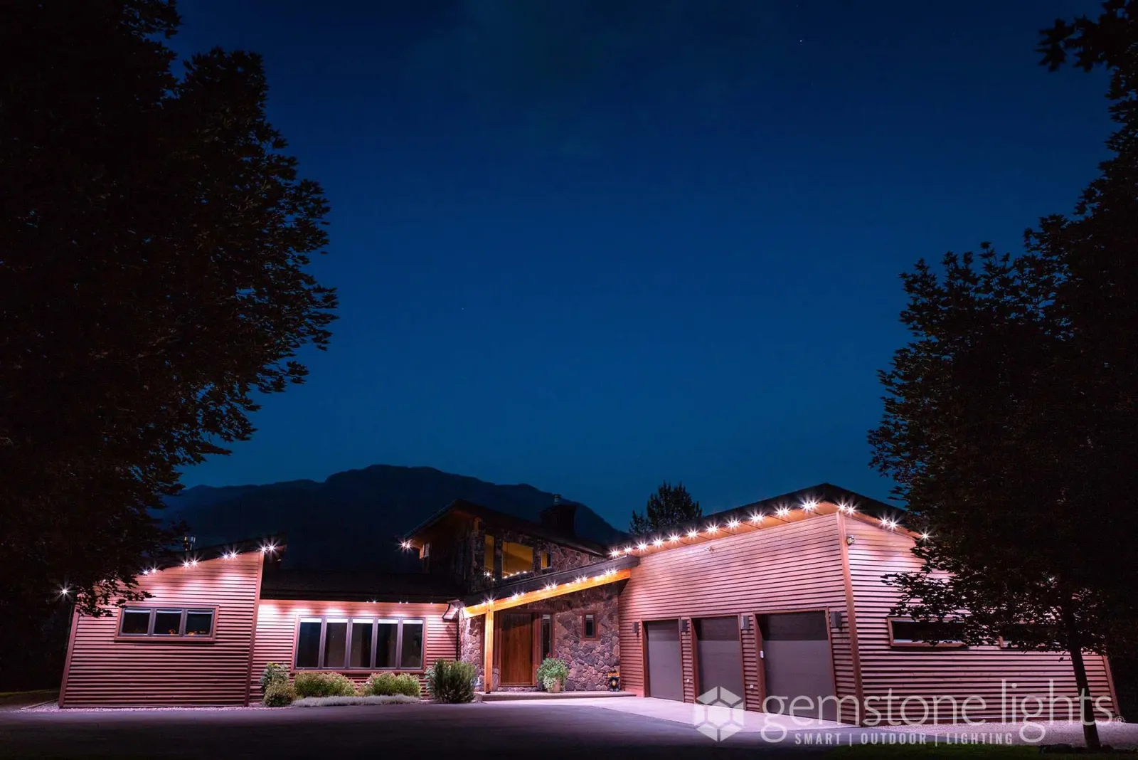 Low ranch-style home surrounded by tall dark trees, its eaves glowing with warm white lights at night