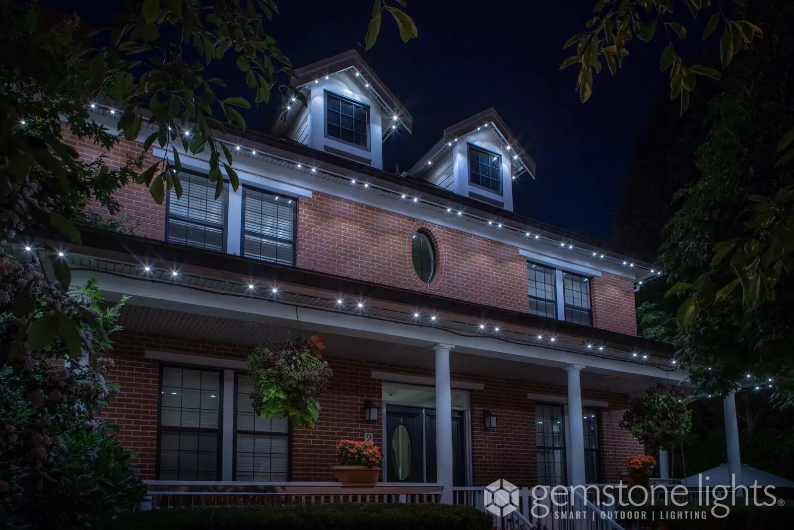 Two-story brick house with a white columned porch, its rooflines and dormers traced in warm white lights at dusk