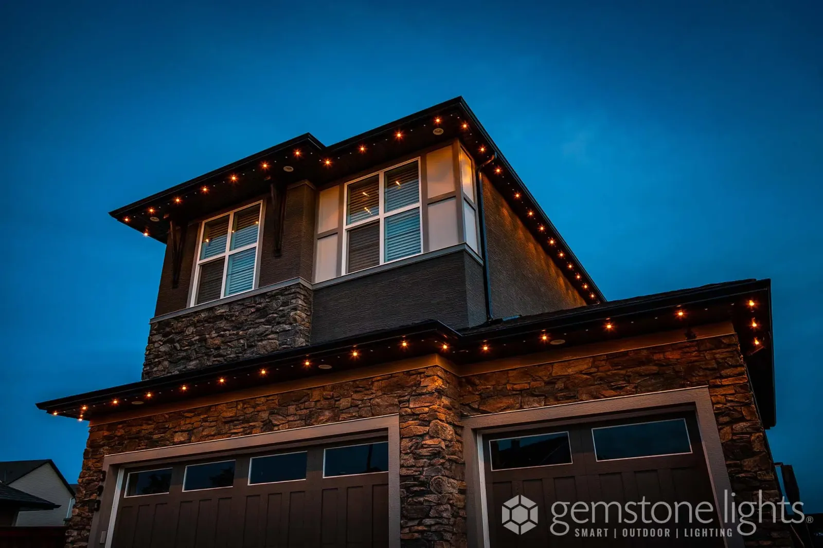Two-story stone and siding home with every roofline traced in warm white lights at dusk